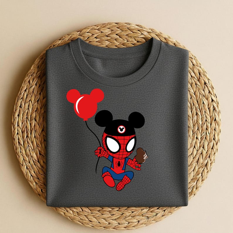 May include: Dark gray t-shirt featuring a cartoon Spider-Man character with Mickey Mouse ears, holding a red balloon and an ice cream cone. The design is in red, blue, and black. The shirt is displayed on a woven, circular placemat.