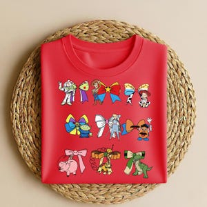 Puede incluir: Una camiseta roja de cuello redondo con lazos de personajes de dibujos animados. Los lazos presentan personajes de la franquicia Toy Story, como Buzz Lightyear y Woody. La camiseta se muestra sobre un salvamanteles redondo tejido.