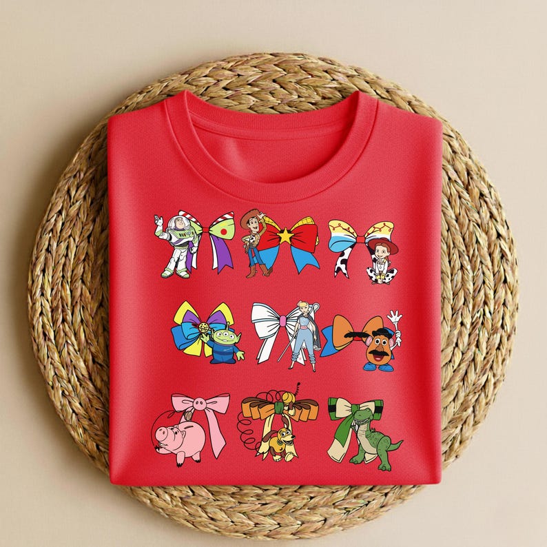 Puede incluir: Una camiseta roja de cuello redondo con lazos de personajes de dibujos animados. Los lazos presentan personajes de la franquicia Toy Story, como Buzz Lightyear y Woody. La camiseta se muestra sobre un salvamanteles redondo tejido.