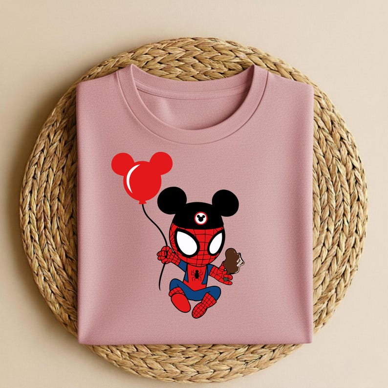 May include: A light pink crew neck t-shirt featuring a cartoon character that is a hybrid of Spider-Man and Mickey Mouse. The character is holding a red balloon and a chocolate ice cream cone. The shirt is folded on a woven mat.