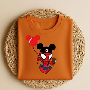 May include: Orange t-shirt featuring a cartoon character that is a hybrid of Spider-Man and Mickey Mouse. The character is holding a red balloon and a chocolate ice cream cone. The shirt is folded on a woven, circular placemat.