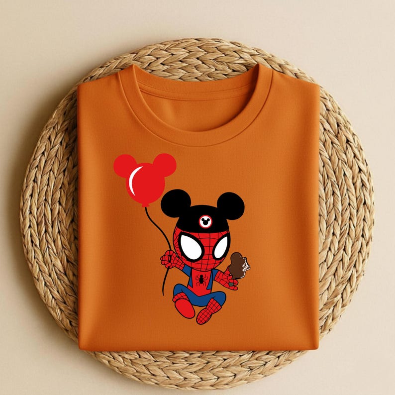 May include: Orange t-shirt featuring a cartoon character that is a hybrid of Spider-Man and Mickey Mouse. The character is holding a red balloon and a chocolate ice cream cone. The shirt is folded on a woven, circular placemat.