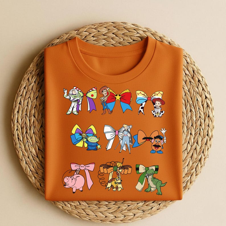 Puede incluir: Camiseta naranja de cuello redondo con dise&ntilde;os de lazos de dibujos animados con personajes. Los lazos est&aacute;n dispuestos en filas e incluyen personajes como Buzz Lightyear, Woody y otros de una popular pel&iacute;cula animada.