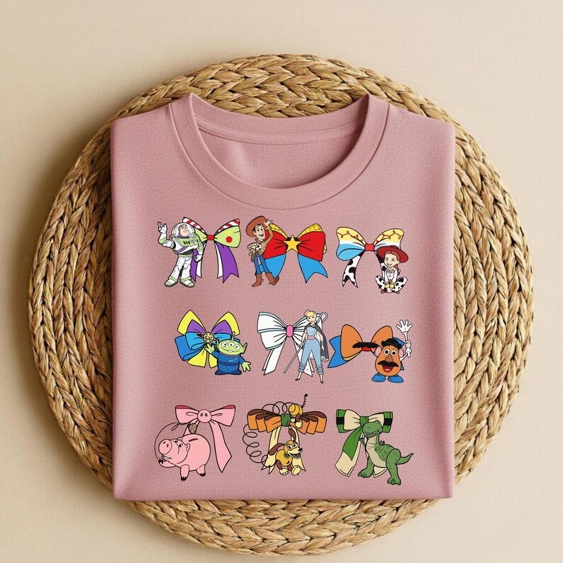 Puede incluir: Una sudadera de color malva con lazos de personajes de dibujos animados. Los lazos presentan personajes como Buzz Lightyear y Woody. La sudadera doblada est&aacute; colocada sobre una estera redonda tejida.