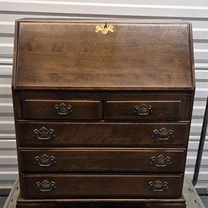 May include: A dark brown wooden secretary desk featuring a slanted drop-front lid with a brass keyhole escutcheon. The piece includes two small upper drawers and three full-width lower drawers, all equipped with matching brass swing-bail handles.