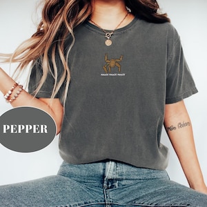 May include: A charcoal gray short-sleeve t-shirt featuring a small brown graphic of a crab-like creature centered on the chest. Below the graphic, the text reads AMAZE AMAZE AMAZE in white capital letters. The shirt has a crew neckline and a relaxed fit.