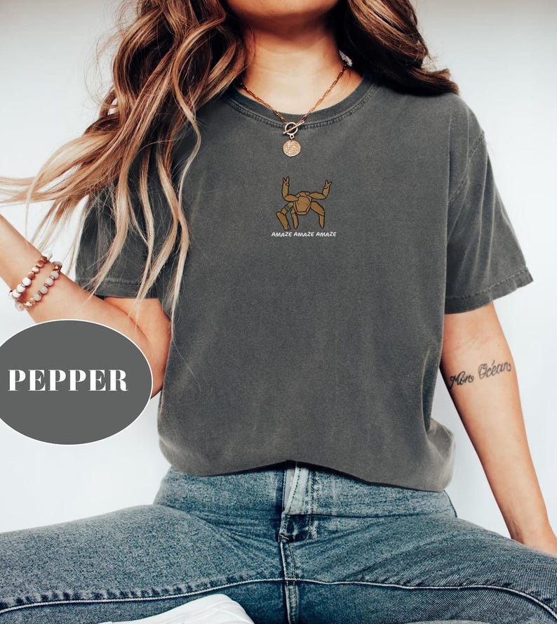 May include: A charcoal gray short-sleeve t-shirt featuring a small brown graphic of a crab-like creature centered on the chest. Below the graphic, the text reads AMAZE AMAZE AMAZE in white capital letters. The shirt has a crew neckline and a relaxed fit.