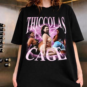May include: A black short-sleeve t-shirt featuring a graphic print with the text THICCOLAS CAGE in large pink letters. The design includes three images of a person with the face of Nicolas Cage superimposed on various bodies against a purple lightning background.