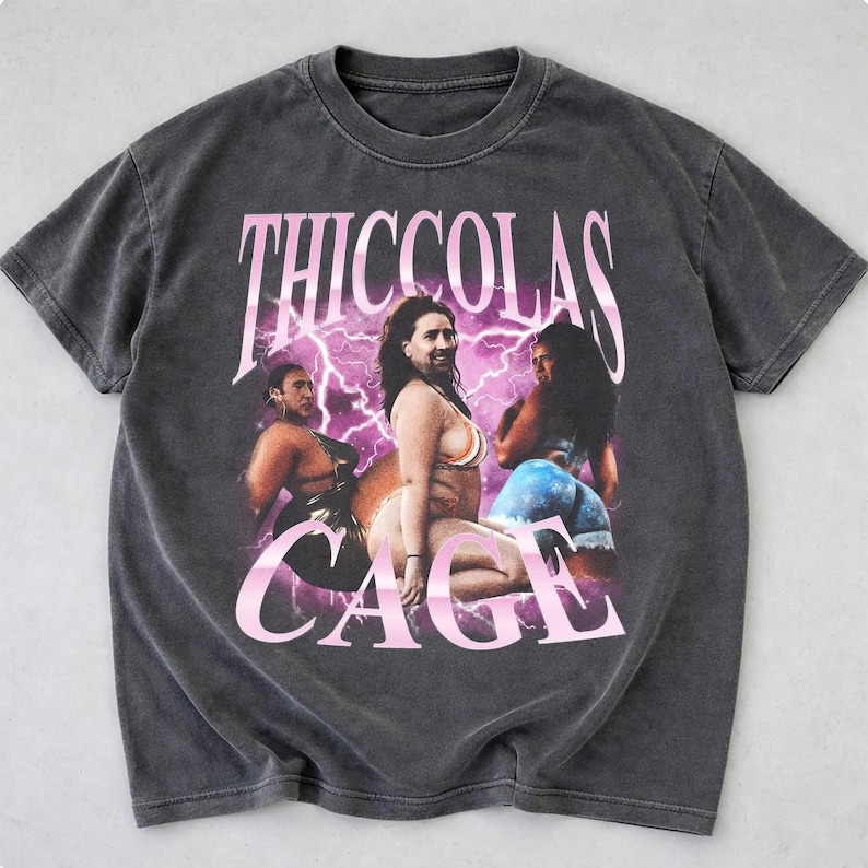 May include: A charcoal gray short-sleeve t-shirt featuring a graphic print with the text THICCOLAS CAGE in a light pink font. The design displays three images of a person with the face of Nicolas Cage superimposed on bodies against a purple lightning background.