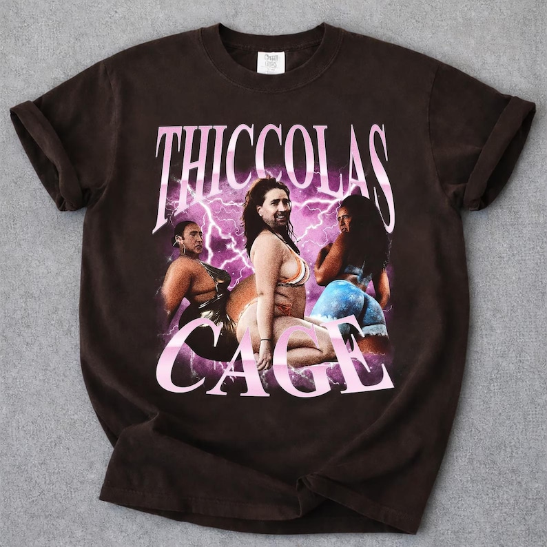 May include: A dark brown short-sleeve t-shirt featuring a graphic print with the text THICCOLAS CAGE in pink. The design includes three images of a person with a beard and long hair, set against a background of bright pink lightning bolts.