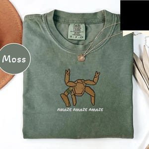 May include: A moss green crew neck t-shirt featuring an embroidered brown spider-like creature with raised legs and the text "AMAZE AMAZE AMAZE" in white. The shirt has a Comfort Colors label at the inner neckline and a small gold-toned pendant necklace.