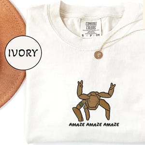 May include: An ivory t-shirt featuring an embroidered brown creature with multiple limbs making peace signs, accompanied by the text "AMAZE AMAZE AMAZE" in black. The shirt has a Comfort Colors label at the neck and is paired with a gold-toned necklace.