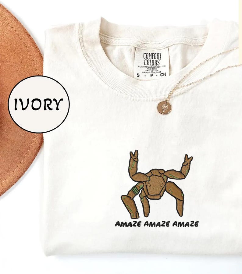 May include: An ivory t-shirt featuring an embroidered brown creature with multiple limbs making peace signs, accompanied by the text "AMAZE AMAZE AMAZE" in black. The shirt has a Comfort Colors label at the neck and is paired with a gold-toned necklace.