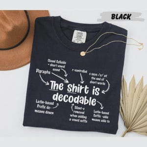 May include: A black short-sleeved t-shirt featuring white text that reads The shirt is decodable, surrounded by arrows pointing to linguistic explanations including Closed Syllable, Digraphs, r-controlled, and Latin-based prefix and suffix definitions.