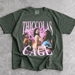 May include: A dark olive green short-sleeve t-shirt featuring a graphic print of three individuals in swimwear. The text THICCOLAS CAGE is printed in large pink letters above the figures, set against a background of purple lightning bolts.