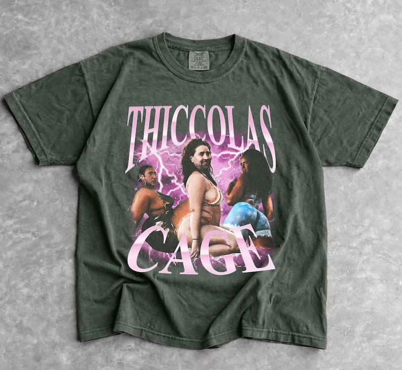 May include: A dark olive green short-sleeve t-shirt featuring a graphic print of three individuals in swimwear. The text THICCOLAS CAGE is printed in large pink letters above the figures, set against a background of purple lightning bolts.