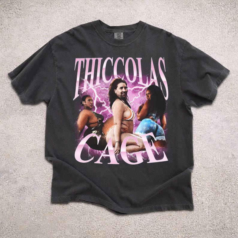 May include: A charcoal gray short-sleeve t-shirt featuring a graphic print of three figures with the face of Nicolas Cage. The design includes pink lightning bolts and the text THICCOLAS CAGE in a bold, stylized pink font across the center.