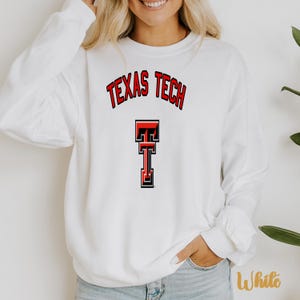 May include: A white long-sleeve crewneck sweatshirt featuring the text TEXAS TECH in a red and black arched font above a red and black double T logo centered on the chest. The garment is made of a solid white fabric with a standard ribbed neckline.