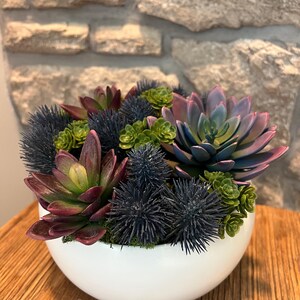 May include: A white, round ceramic bowl filled with a variety of artificial succulents. The arrangement includes green, purple, and blue-green succulents, along with dark blue spiky accents. The bowl sits on a wooden surface.