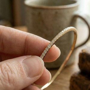 Puede incluir: Un brazalete de oro con una fila de pequeños diamantes brillantes. La pulsera está sujeta por una mano, mostrando el diseño delicado y la artesanía. El fondo incluye una taza y una superficie de madera.