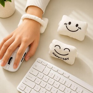 Pode incluir: Um conjunto de apoios de pulso de pelúcia branca para computador, feitos de tecido macio com rostos sorridentes bordados. Um apoio retangular é usado no pulso, enquanto outros dois estão próximos, um deles com o texto HAPPY DAY bordado em preto.