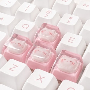 May include: A mechanical keyboard featuring white keys with light pink lettering and four custom artisan keycaps. Each artisan keycap is made of clear resin encasing a small, sculpted white cat figure with pink facial details and a pink base.