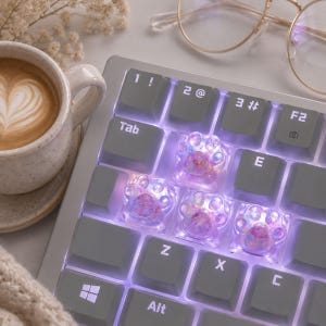 May include: A mechanical keyboard featuring custom translucent cat paw keycaps with iridescent glitter accents. The keycaps are illuminated by a soft purple backlight, replacing standard keys. The keyboard has grey keys with white lettering.