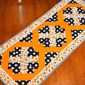 May include: A quilted table runner with a patchwork design. It features orange, navy blue, and white fabrics. The runner has a repeating pattern of navy blue polka dots and orange squares. The edges are finished with a scalloped trim and a black and white striped border.