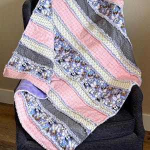 May include: A handmade rag quilt with a patchwork design. The quilt features alternating stripes of pink, gray, and patterned fabric with bunnies. The quilt is draped over a dark gray chair.