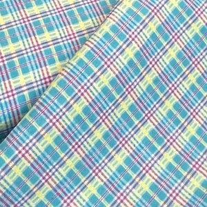May include: Close-up of a fabric with a plaid pattern. The fabric features a repeating pattern of intersecting lines in teal, yellow, white, and purple. The fabric is folded over, showing the pattern from different angles.