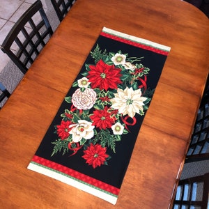 May include: A black table runner with a floral design featuring red and white poinsettias, green leaves, and white ribbon accents. The runner has a green and red border.
