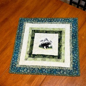 May include: A square quilted placemat with a bear and mountain design. The placemat features a central square with a black bear silhouette, mountains, and trees. The design is surrounded by green, white, and blue patterned borders.