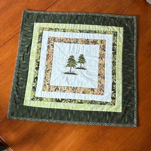Peut inclure: Nappe carrée matelassée avec un motif brodé de deux arbres verts au centre. Elle présente des carrés concentriques de tissu blanc, vert et à motifs, encadrés par une bordure vert foncé. Le design a un thème inspiré de la nature.
