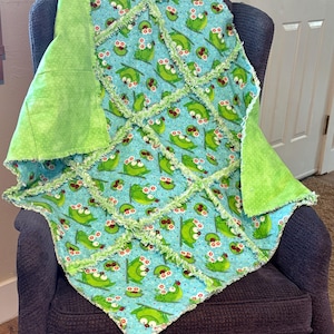 Könnte beinhalten: Eine handgefertigte Babydecke mit einem skurrilen Froschmuster auf hellblauem Hintergrund. Die Decke hat ein Patchwork-Design mit grünen Akzenten und fühlt sich weich und strukturiert an. Die Kanten sind mit einer Rüschenborte versehen.