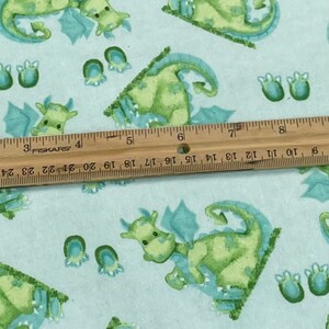 May include: Light blue fabric with a repeating pattern of green dragons with blue wings and blue footprints. The fabric is measured with a ruler.