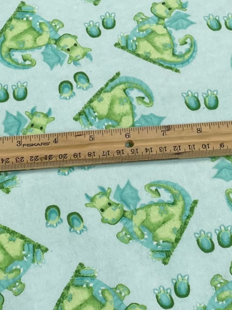 May include: Light blue fabric with a repeating pattern of green dragons with blue wings and blue footprints. The fabric is measured with a ruler.