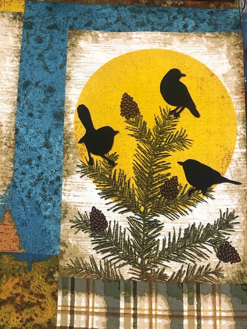 May include: A fabric panel featuring a nature scene with a yellow sun, black bird silhouettes, pine needles, and pine cones. The background includes blue, green, and brown textured patterns and a plaid design.