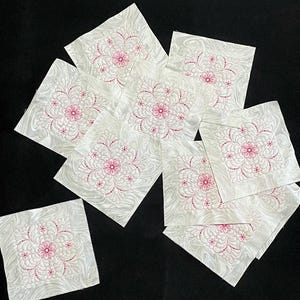 Pode incluir: Dez quadrados de tecido branco com bordados florais rosa. Os quadrados estão dispostos em um padrão aleatório sobre um fundo preto.