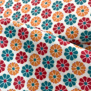 May include: White fabric with a repeating pattern of stylized flowers in red, teal, and orange. The flowers have a central orange circle with petals in the other colors. The fabric is folded, showing the pattern at different angles.