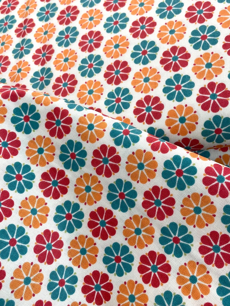 May include: White fabric with a repeating pattern of stylized flowers in red, teal, and orange. The flowers have a central orange circle with petals in the other colors. The fabric is folded, showing the pattern at different angles.
