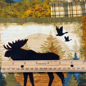 May include: A fabric panel featuring a forest scene with a moose silhouette, flying geese, and pine trees. The design incorporates a plaid pattern and a ruler with inch measurements. Colors include green, blue, brown, and tan.