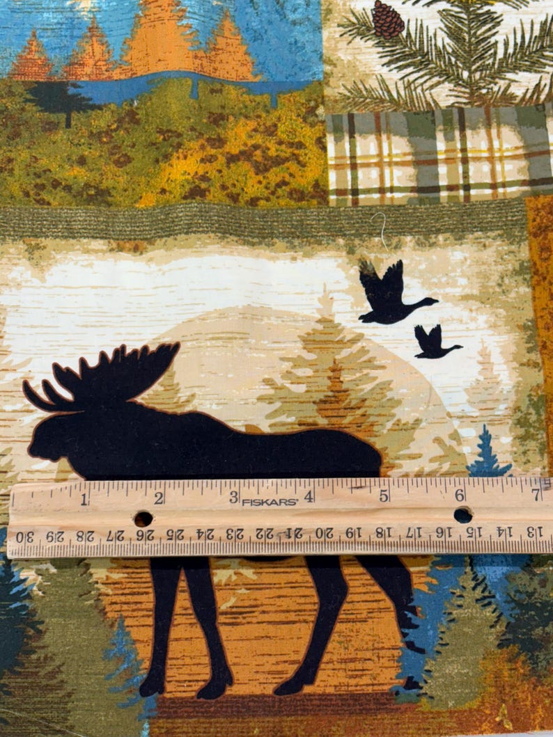 May include: A fabric panel featuring a forest scene with a moose silhouette, flying geese, and pine trees. The design incorporates a plaid pattern and a ruler with inch measurements. Colors include green, blue, brown, and tan.