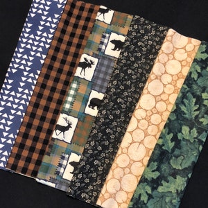 May include: A collection of fabric strips with various patterns. Designs include blue triangles, brown plaid, moose and bear silhouettes, floral, wood grain, and green leaves. The fabrics are arranged on a black surface.