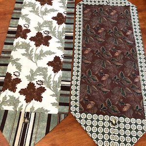 May include: Two table runners with a brown and green color scheme. One runner features a floral pattern with brown and green leaves and flowers on a white background. The other runner features a brown and green leaf pattern on a brown background. Both runners have a green and brown border.