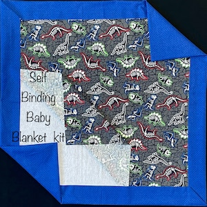 Op de afbeelding: Een blauwe en witte babydekenset met een dinosaurus skeletprint. De set bevat stof voor de deken en de binding. De tekst "Self Binding Baby Blanket Kit" is zichtbaar op de stof.