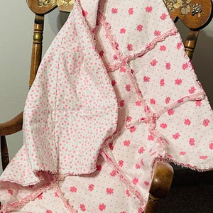 Pode incluir: Um cobertor de bebê em patchwork rosa e branco com uma borda franjada. O cobertor é feito de tecido de algodão macio e apresenta uma estampa floral.