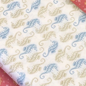 Op de afbeelding: Witte stof met een herhalend patroon van blauwe en beige zeepaardjes. De stof heeft een subtiele textuur.