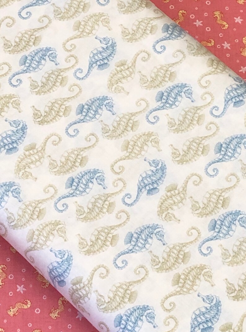Op de afbeelding: Witte stof met een herhalend patroon van blauwe en beige zeepaardjes. De stof heeft een subtiele textuur.