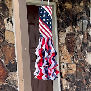 May include: A hanging windsock featuring a cylindrical top section patterned with the American flag design of white stars on a blue field and red and white stripes. Below the cylinder are numerous long, curling ribbons in red, white, and blue.