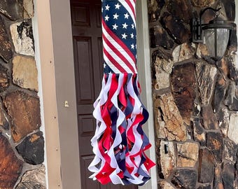 May include: A hanging windsock featuring a cylindrical top section patterned with the American flag design of white stars on a blue field and red and white stripes. Below the cylinder are numerous long, curling ribbons in red, white, and blue.
